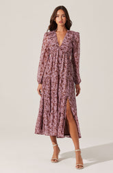 A woman is wearing a long, mauve-colored, patterned dress with long sleeves and puffed shoulders. The dress has a deep V-neckline and features a high slit on one side, revealing her leg. She is also wearing strappy high-heeled sandals.